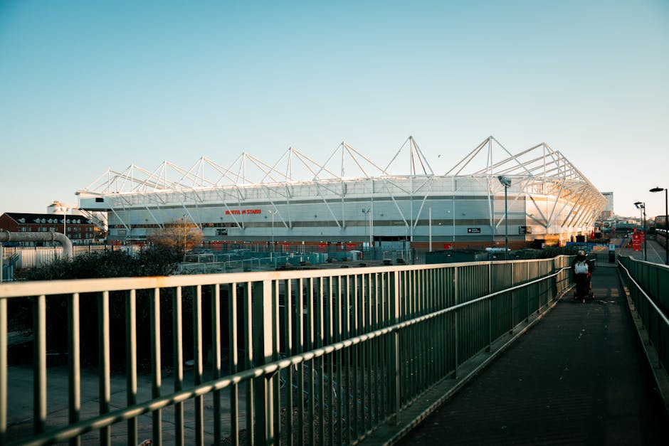 A modern football stadium with a white exterior and a distinctive, geometric roof structure supported by white steel trusses, situated behind a green metal railing on a pedestrian pathway. The pathway