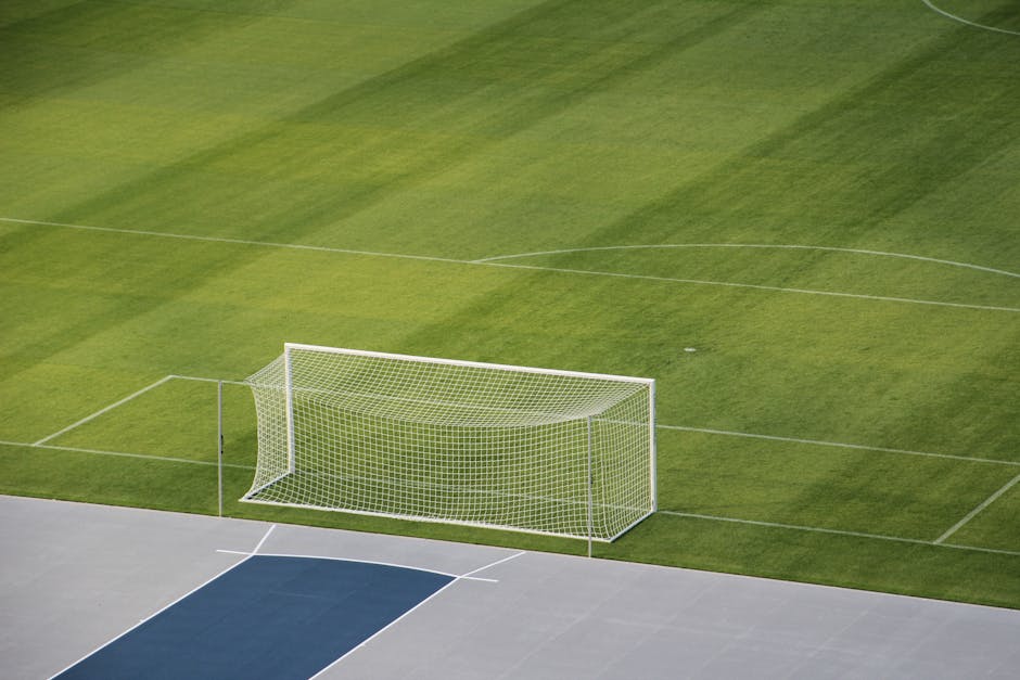 A modern football stadium with a white exterior and a distinctive, geometric roof structure supported by white steel trusses, situated behind a green metal railing on a pedestrian pathway. The pathway